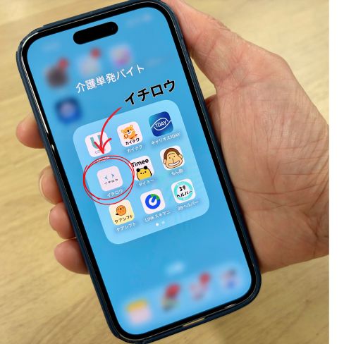 かくと（長田哲也）が持つスマホにダウンロードされた9つの介護単発バイトアプリの写真。わかりやすく丸印と矢印でイチロウアプリのみ指している画像。